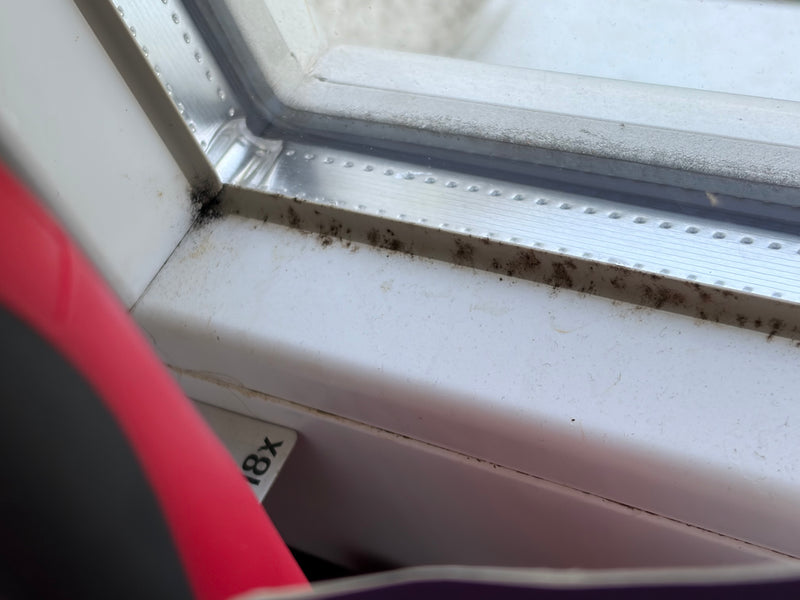 De foto toont donkere schimmelgroei in de hoek van een wit raamkozijn. Dit is typische zwarte schimmel.