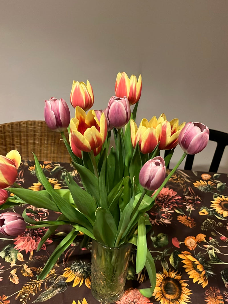 Een kleurrijk boeket tuintulpen staat in een glazen vaas op een gedessineerde tafel. De bloemen tonen verschillende kleuren zoals rood, geel en roze.