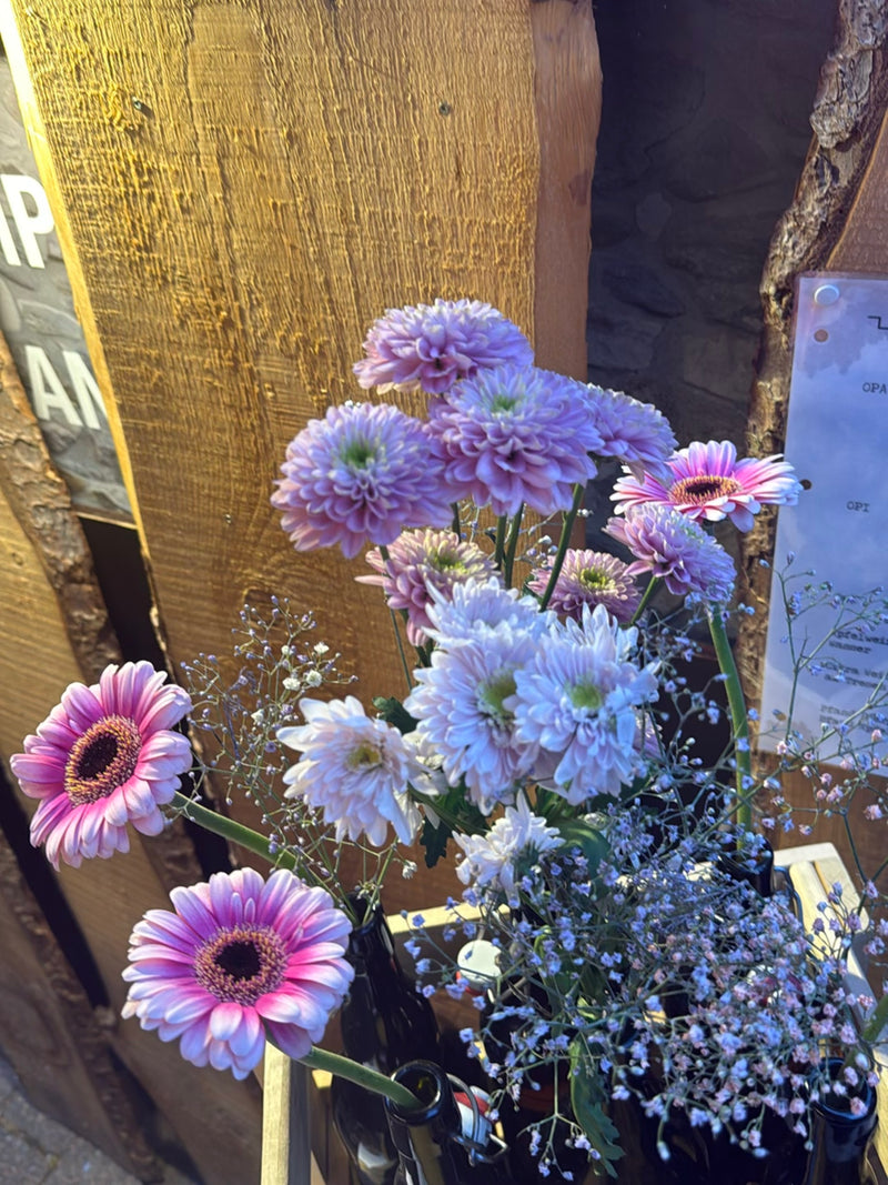Verschillende roze gerberabloemen staan samen met andere bloemen in flessen voor een houten muur.