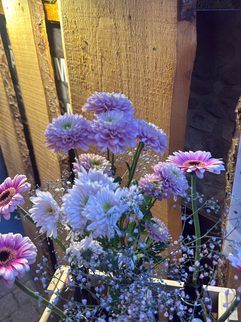 Te zien is een kleurrijk boeket bloemen met gerbera's en andere bloesems voor een houten wand. De planten staan in bakken.