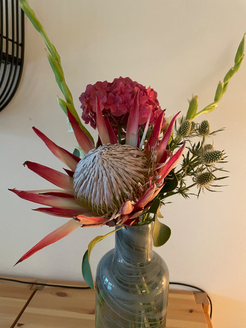 Een grote bloem van de koningsprotea (Protea cynaroides) is zichtbaar als onderdeel van een kleurrijk boeket in een glazen vaas. De opvallende, puntige schut...