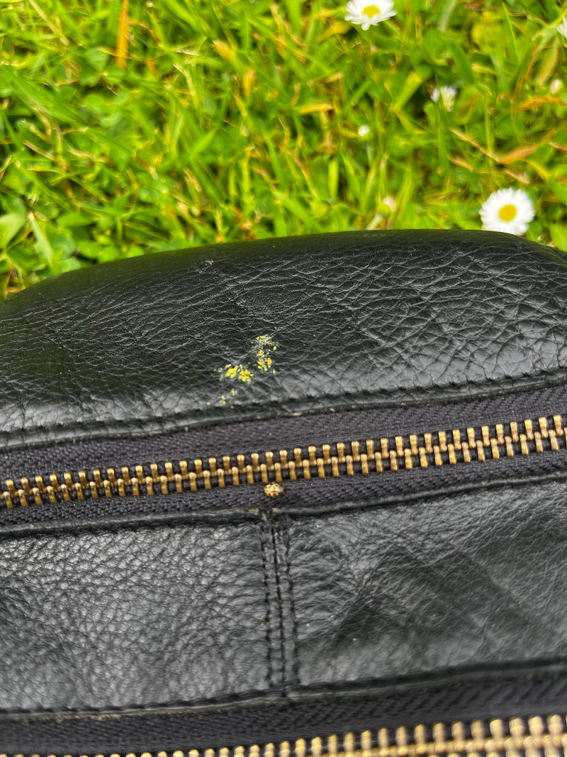 Een zestienvlekkige lieveheersbeestje zit op de rits van een zwarte lederen tas. Op de achtergrond is een groen weiland met margrietjes te zien.